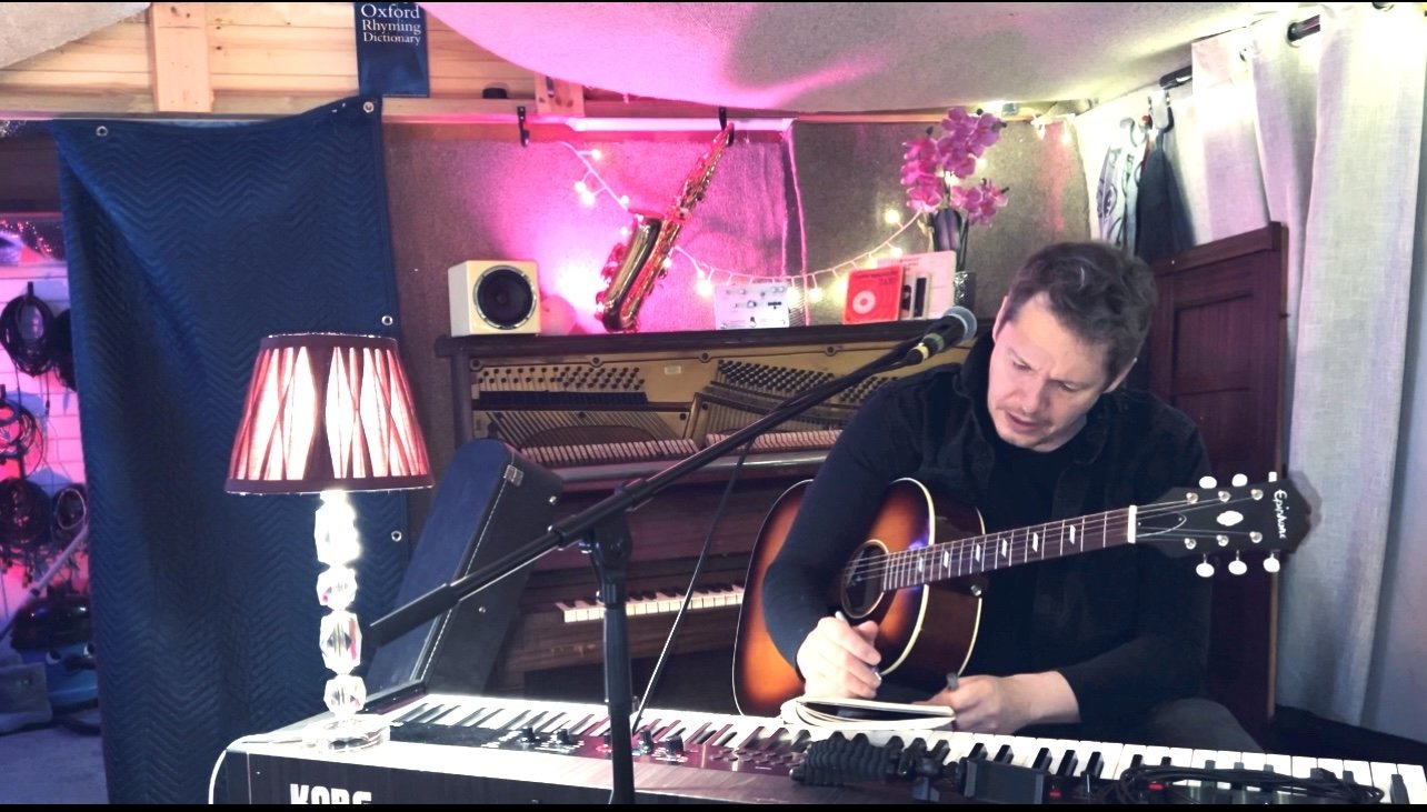 Rich Chance playing guitar in his home studio, piano and saxophone in the background