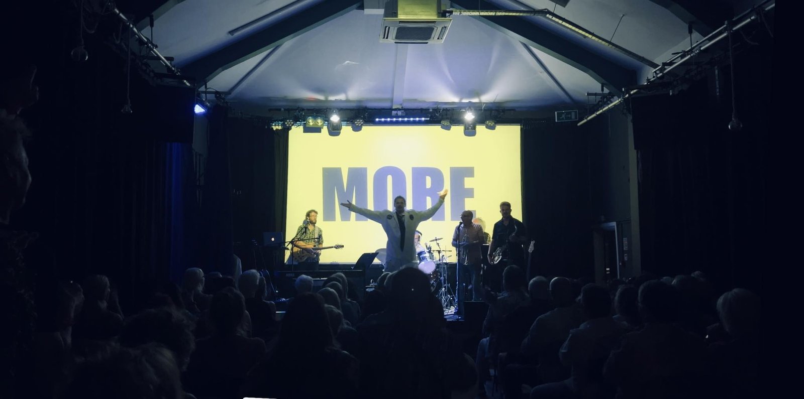 Rich Chance performing live, arms outstretched in front of a screen reading MORE